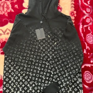 Svart Louis Vuitton hoodie med monogram - Svart hoodie från Louis Vuitton med klassiskt vitt monogrammönster över hela tröjan. Tröjan har huva med snörning och ribbade muddar. Materialet är mjukt och bekvämt, perfekt för dig som vill sticka ut med en lyxig streetstyle-look.