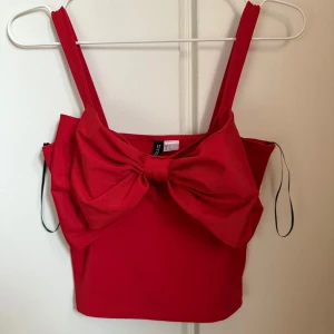 Röd croppad topp med rosett från H&M - Säljer en röd croppad topp från H&M Divided med breda axelband och en stor, iögonfallande rosett framtill. Toppen har en tight passform och är tillverkad i mjuk polyester. Perfekt för dig som vill sticka ut med en färgstark och trendig look. Använd en gång.