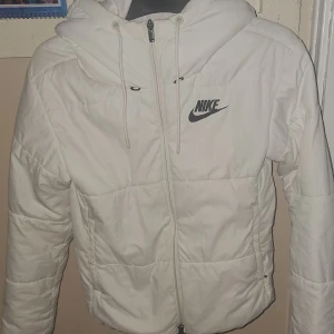 Vit pufferjacka från Nike med huva - Snygg vit pufferjacka från Nike med svart logga på bröstet. Jackan har huva med snörning, dragkedja framtill och quiltad design. Perfekt för kyliga dagar och ger en sportig vibe.