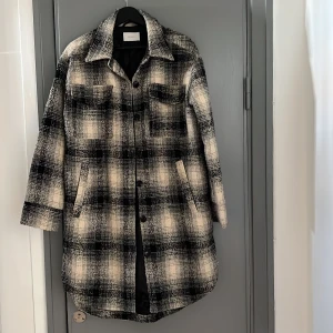 Rutig oversize skjortjacka från Neo Noir - Lång, rutig oversize skjortjacka från Neo Noir i svart,grå och beige. Jackan har klassisk krage, knäppning framtill, två bröstfickor med lock och två sidofickor. Perfekt lager-på-lager-plagg med avslappnad passform och lång ärm.