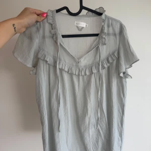 Mint grön blus med volang och knyt - Supersöt ljusgrå blus från Amber & Cloud med volangdetaljer längs v-ringningen och axlarna. Blusen har korta ärmar och knytband vid halsen för extra detalj. Luftigt material som känns skönt mot huden och passar perfekt till jeans.