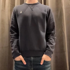 Mörkblå crewneck från Polo Ralph Lauren - En stilren mörkblå crewneck från Polo Ralph Lauren med klassisk rund halsringning och broderad vit logga på bröstet. Tröjan är i mjuk bomull och har långa ärmar samt ribbade muddar vid ärmslut och nederkant. Perfekt för en clean och avslappnad look.