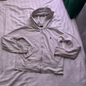 Vit hoodie från Polo Ralph Lauren - Vit hoodie från Polo Ralph Lauren med dragkedja framtill, två fickor och klassisk broderad logga på bröstet. Tröjan har huva och långa ärmar, tillverkad i mjuk bomull för en chill och bekväm stil. liten fläck på tröjan men inget man märker.
