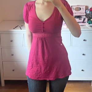 Mörkrosa babydoll blus med v-ringning - Supersöt mörkrosa blus med v-ringning, korta puffärmar och knappar framtill. Modellen är lite längre och har markerad midja som ger en snygg siluett. Perfekt att styla med jeans eller kjol för en trendig look.