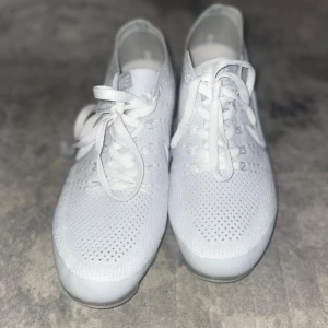 Nike Vapormax Flyknit vita sneakers - Säljer ett par fräscha Nike Vapormax Flyknit sneakers i helvitt. Skorna har en stickad ovandel i syntetmaterial, klassisk snörning och genomskinlig sula med den ikoniska Vapormax-dämpningen. Perfekta för dig som gillar stilrena och sportiga sneakers.