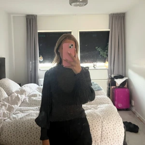 Glittrig svart topp med puffärm - Säljer en svart glittrig topp med höga puffärmar och smal passform. Toppen har lång ärm och en lite högre hals, vilket ger en trendig och cool look. Perfekt för dig som vill sticka ut med lite extra glitter.