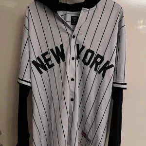 Randig baseballtröja med huva från FSBN - Säljer en oversized vit och svart randig baseballtröja från FSBN med trycket NEW YORK på bröstet. Tröjan har svarta detaljer, knappar framtill, svart huva och långa svarta ärmar under de korta. Märke och patch nertill med USA 1992.