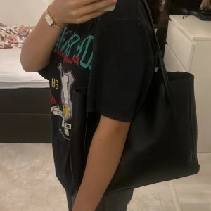 Svart axelväska i skinnimitation - Stilren svart axelväska med långa handtag och rymlig insida. Väskan är i slät skinnimitation och har en enkel, klassisk design utan synliga detaljer eller mönster. Perfekt storlek för att få plats med allt du behöver under dagen. Bredden är 34cm och längden är 28cm.