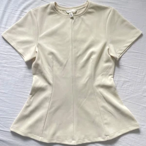 Ljusgul figursydd topp från H&M - Säljer en stilren beige topp från H&M med korta ärmar och rund halsringning. Toppen är figursydd med sömmar och är perfekt att matcha med jeans eller kjol för en clean look.