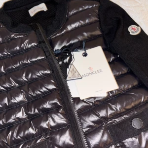Svart dunväst från Moncler - Snygg svart dunväst från Moncler med quiltad design och glansig finish. Västen har dragkedja framtill, ribbade detaljer och klassisk Moncler-logga på bröstet. Perfekt för lager-på-lager och en stilren look.
