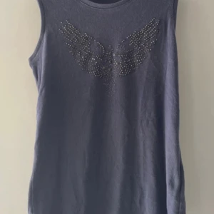 Grå ärmlös topp med nitar från MRK Adam - Snygg grå ärmlös linnetopp från MRK Adam med coolt nitmönster i form av vingar framtill. Toppen har rund halsringning och är gjord i mjuk bomull. Perfekt för dig som gillar edgy detaljer och vill sticka ut lite extra.