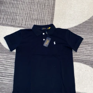 Mörkblå Polo Ralph Lauren t-shirt - Klassisk mörkblå t-shirt från Polo Ralph Lauren med korta ärmar och vit broderad logga på bröstet. T-shirten har krage och knappar framtill, tillverkad i 100% bomull för en skön och stilren look. Åker du på semester under dom här kommande kalla tider är denna tröja perfekt för dig. Pris kan diskuteras vid snabbaffär!!