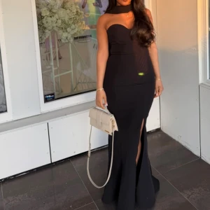 Svart axelbandslös långklänning - Supersnygg svart axelbandslös långklänning med figurnära passform och en elegant slits framtill. Klänningen har en hjärtformad byst och är perfekt för dig som vill sticka ut med en stilren och tidlös look.