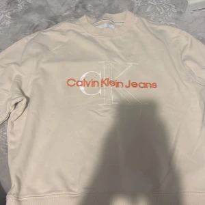 Beige sweatshirt från Calvin Klein Jeans - Snygg beige sweatshirt från Calvin Klein Jeans med broderad logga i vitt och orange framtill. Tröjan har rund halsringning, ribbade muddar och är tillverkad i mjuk bomullsmix. Perfekt för en avslappnad och trendig look.