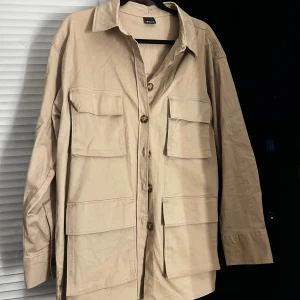 Beige overshirt från Gina Tricot - Säljer en beige overshirt från Gina Tricot i storlek L. Skjortan har fyra stora fickor framtill, klassisk krage och knäppning med snygga knappar. Tillverkad i bomull och har en loose passform med långa ärmar. Perfekt för lager-på-lager-stil.
