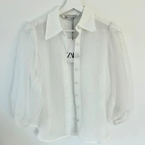 Vit transparent skjorta från Zara - Säljer en vit transparent skjorta från Zara med diskreta prickar i tyget. Skjortan har klassisk krage, knappar framtill och puffiga trekvartsärmar med resår vid ärmslut. Perfekt för en trendig och luftig look.