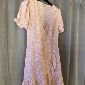 Ljusrosa omlottklänning från NLY Trend - Supersöt ljusrosa kortklänning från NLY Trend med vitt blommigt mönster. Omlottmodell med knyt i midjan, puffiga korta ärmar och volangkant nertill. Perfekt för sommarens alla tillfällen och ger en romantisk vibe.ena snöret är borta men inget som syns