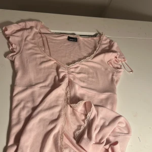 Ljusrosa topp med spets från Gina Tricot - Supersöt ljusrosa topp från Gina Tricot med korta ärmar, v-ringning och fina spetsdetaljer längs kanterna. Toppen har rynk framtill och är gjord i mjuk bomull. Perfekt för dig som gillar romantisk och feminin stil.