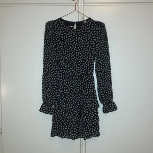 Svart blommig klänning H&M Divided - Supersöt svart kortklänning från H&M Divided med vitt blommönster. Klänningen har långa ärmar med volangdetalj vid ärmslut och en lätt rynkad kjol. Strl 32. Knappt använd, väldigt bra skick!!!