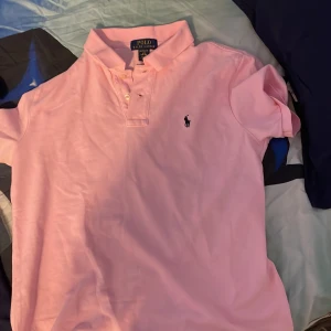  Äkta Rosa pikétröja från Polo Ralph Lauren - Snygg rosa pikétröja från Polo Ralph Lauren med klassisk krage och knappar framtill. Tröjan har korta ärmar och den ikoniska broderade loggan på bröstet. Perfekt för en clean och stilren look.