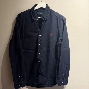 Ralph Lauren skjorta  - Säljer en mörkblå skjorta från Ralph Lauren i slim fit-modell. Skjortan har klassisk krage, vita knappar. Tillverkad i mjuk bomull och har långa ärmar med knapp vid manschetten.