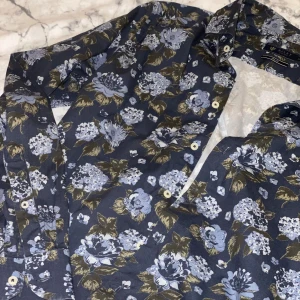 Blommig skjorta från Riley slim fit - Snygg mörkblå skjorta från Riley med blommigt mönster i ljusblått och grönt. Slim fit-modell med klassisk krage och knappar framtill. Perfekt för dig som vill sticka ut med en cool och fräsch look. Materialet känns lätt och skönt mot huden. Finns extra knappar. 100% Cotton 😊