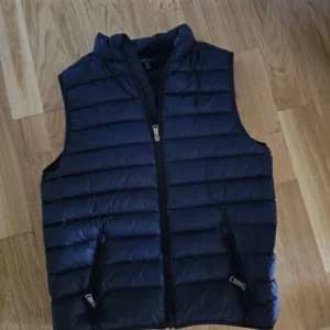 Blå dunväst från Ralph Lauren - Säljer en snygg blå dunväst från Ralph Lauren. Västen har en dragkedja framtill och två praktiska fickor med dragkedjor. Perfekt för kyligare dagar när du vill hålla stilen. Säljer pga att den är för liten. Bra skick utöver de 2 lappade hålen ni ser i bilderna.