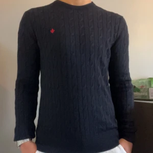 Mörkblå kabelstickad tröja Morris XS - Kabelstickad mörkblå tröja från Morris i 100% merinoull. Klassisk rund halsringning och diskret röd logga broderad på bröstet. Perfekt för dig som gillar stilrena och tidlösa plagg med en snygg twist.