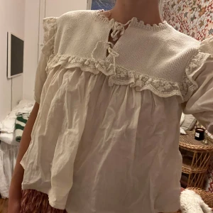 Vit romantisk blus med spets och knyt - Superfin vit blus med stickad överdel och volangkant i spets. Blusen har korta puffärmar och knytning fram vid halsen. Den är luftig och har en söt, romantisk känsla med detaljer i både bomull och stickat material.