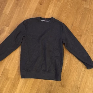 Mörkgrå sweatshirt från Tommy Hilfiger S - Mörkgrå sweatshirt från Tommy Hilfiger med klassisk rund hals och diskret broderad logga på bröstet. Tröjan har ribbade muddar vid ärmslut och nederkant samt snygga detaljer i rött, vitt och blått vid kragen. Perfekt för en stilren och avslappnad look. Storlek S, säljer för den inte passar mig längre.