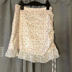 Vit blommig kortkjol med volang - Supersöt vit kortkjol med små blommor i rosa och orange, volangkant i spets och snörning på sidan. Kjolen har en lätt glansig yta och är perfekt för vår och sommar. Sitter snyggt med en normal passform och har en romantisk vibe.