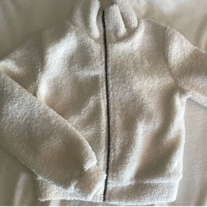 Vit teddyjacka med dragkedja - Supermjuk och fluffig vit teddyjacka med hög krage och hel dragkedja framtill. Jackan har långärmade ärmar och är fodrad på insidan för extra komfort. Perfekt för kyliga dagar när du vill vara både varm och snygg.