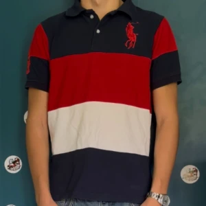 Ralph Lauren randig pikétröja - Säljer en klassisk pikétröja från Ralph Lauren med breda ränder i marinblått, rött och vitt. Tröjan har korta ärmar, krage och knappar framtill samt stor röd logga på bröstet och siffran 3 på ärmen. Perfekt för en sportig och stilren look.
