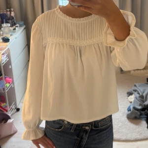 Vit blus med volang och spetsdetalj - Supersöt vit blus med långärmade volangslut och rynkad överdel. Blusen har fina spetsdetaljer och lätt struktur i tyget. Perfekt till jeans för en fräsch look. Hög halsringning och avslappnad passform.
