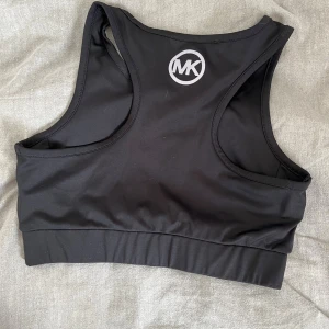 Svart linnetopp från Michael Kors - Sportig svart linnetopp från Michael Kors med rund halsringning och brett band nedtill. Toppen har en stilren design med MK-logga framtill och är tillverkad i ett stretchigt syntetmaterial som passar perfekt för träning.