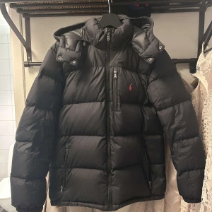 Polo Ralph Lauren Jacka - Svart pufferjacka från Polo Ralph Lauren med huva och dragkedja framtill. Jackan. Perfekt för kalla dagar och riktigt snygg streetstil. Säljer den eftersom jag växt ur den, enda felet med den är lagning på ryggen men syns knappast därav billagare pris, ett riktigt kap! Den passar dig som är 170-175 cm. 