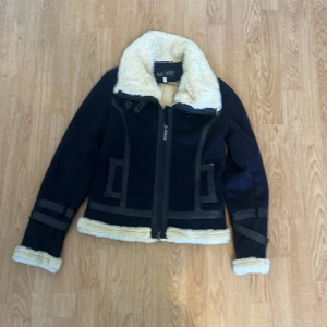 Svart pälsjacka från Armani Jeans - Snygg svart pälsjacka från Armani Jeans med krage och detaljer i beige fuskpäls. Jackan har dragkedja framtill, dekorativa sömmar och coola spännen vid ärmsluten. Perfekt för kalla dagar när du vill vara både varm och stilren.