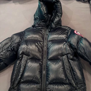 Svart dunjacka från Canada Goose - Säljer en svart, glansig dunjacka från Canada Goose med stor huva och klassisk logga på ärmen. Jackan har dragkedja framtill, två sidofickor med dragkedja och breda quiltade paneler, samt en stor inne ficka och två nät hållare på instan. Insidan har remmar med Canada Goose-tryck för att bära jackan över axeln. Helt oanvänd, för fler bilder samt frågor osv kom pm, helt äkta köpt från Canada Goose. Byten funkar också!