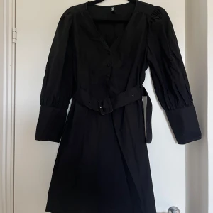 Svart klänning med bälte från Pieces - Svart klänning från Pieces med v-ringning och långa puffärmar. Klänningen har knappar framtill och ett matchande bälte i midjan som ger en snygg siluett. 