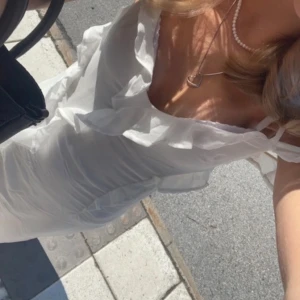 Vit långklänning med volang och knyt - Supersöt långklänning i vit, tunn och flowy polyester. Klänningen har smala axelband, v-ringning, volangdetaljer längs hela framsidan och knyt i ryggen. Perfekt för sommarens alla tillfällen när du vill känna dig extra fin. Den är endast använd en gång på skolavslutning, därav nytt skick och pris! 🌸