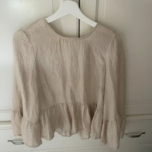 Beige volangblus i bomull - Säljer en ljusbeige blus med volangdetaljer och lång ärm. Blusen är tillverkad i ett tunt, strukturerat bomullstyg och har en avslappnad passform. Perfekt för dig som gillar en luftig och romantisk stil. Köpt på Åhléns!