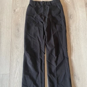 Svarta kostymbyxor från Lindex - Svarta kostymbyxor från Lindex i klassisk rak modell. Byxorna har normal midja, hällor för bälte och stängs med knapp och dragkedja. Snyggt skräddade med pressveck och tillverkade i ett mjukt bomullsmaterial. Byxorna är idagsläget lite skrynkliga men går lätt att åtgärda med ett strykjärn :)