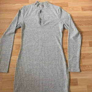 Ny grå ribbad långärmad klänning - Snygg grå ribbad klänning med lång ärm och hög krage. Klänningen har en cool keyhole-detalj framtill och är figurnära i modellen. Perfekt för dig som gillar enkel men trendig stil.