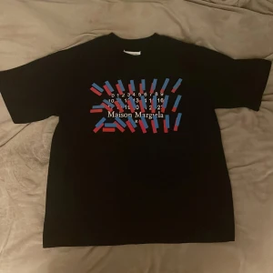 Maison Margiela t-shirt - Svart t-shirt från Maison Margiela med en fet grafiskt print i rött och blått på bröstet och med deras vita logga. Klassisk rund hals och korta ärm. Passformen är loose och t-shirten har en clean look med diskreta vita stygn bak i nacken. Fick den av min brorsa men jag tycker den är för baggy för min del