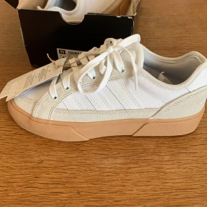 Adidas Court TNS Premiere sneakers - Fräscha Adidas Court TNS Premiere sneakers i vitt med beige detaljer och gummisula. Skorna har klassiska tre ränder på sidan, snörning och en mix av mocka och textil. Perfekta för dig som gillar stilrena och sportiga vibes.