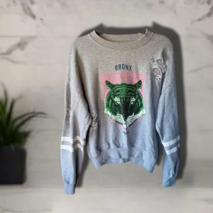 Grå sweatshirt med tigertryck Gina Tricot - Grå sweatshirt från Gina Tricot med stort tigertryck i grönt och rosa på bröstet och texten Bronx New York. Tröjan har slitna detaljer på ärmar och axel samt vita ränder på ärmarna.