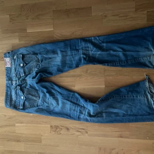 True Religion bootcut jeans blå, stl 29 - Säljer ett par blå bootcut jeans från True Religion med slitna detaljer och trasiga partier framtill. Klassisk femficksmodell, orange sömmar och ikoniska bakfickor. Jeansen är tillverkade i USA och har en cool, vintage vibe. Perfekta för dig som gillar en edgy stil.