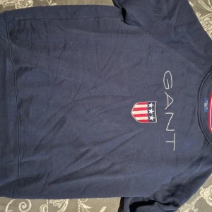Mörkblå sweatshirt från GANT - Snygg mörkblå sweatshirt från GANT med stort tryck och logga på bröstet. Tröjan har klassisk passform och är tillverkad i mjuk bomull. Perfekt för dig som gillar stilrena och sportiga plagg med amerikansk vibe. Lite mindre passform en large skulle jag säga. Pris kan diskuteras. 