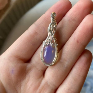 Silverberlock med opalit - Unik handgjord halsbandspendel med en oval, lila sten som skimrar i ljuset. Infattningen är i silverfärgad metall med snygga snirkliga detaljer och tvinnad kant. Perfekt för dig som gillar boho eller magisk vibe. Stenen skiftar mellan lila och vit beroende på ljus.