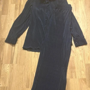 Mörkblå plisse set - Säljer en mörkblå skjorta med byxor till i plisserat tyg och klassisk krage. Skjortan har långa ärmar, knappar framtill och en loose fit. Byxorna är lösa och stretchiga i midjan. Glansigt och mjukt tyg perfekt till en chill men ändå stilren look.